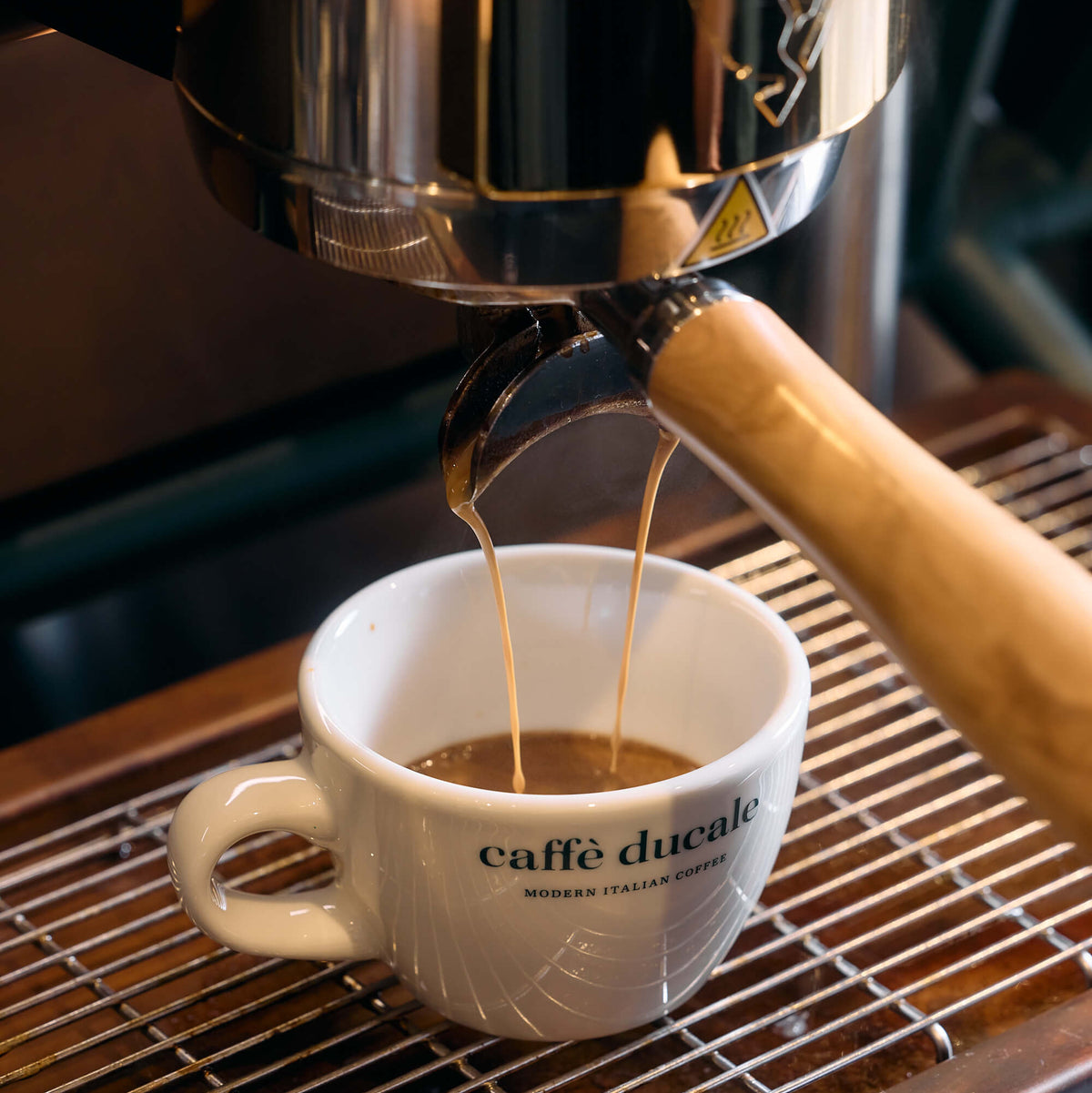 A close-up of espresso being poured from an automatic coffee machine into a white ceramic cup printed with the brand name 'caffè ducale'.